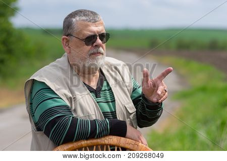 Outdoor portrait of senior man in black sunglasses leant on a wicker chair back and gesticulating