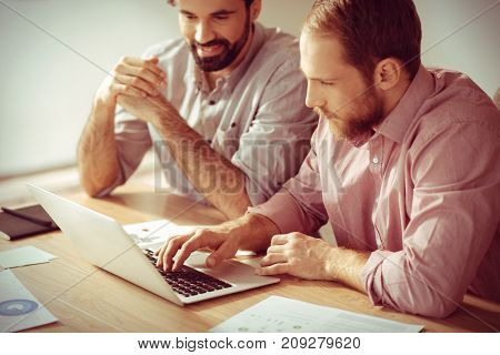 Entering information. Serious smart handsome man sitting in front of the laptop screen with his colleague and pressing buttons on the keyboard while entering information