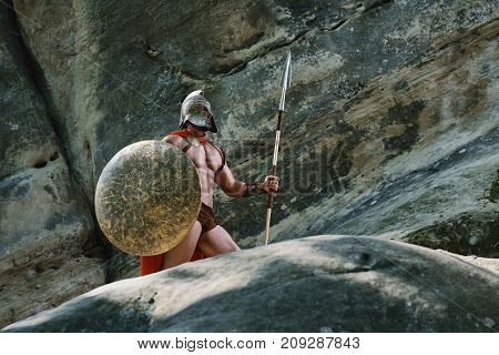 Strong muscular medieval warrior with sexy body and perfect abs wearing a helmet holding a spear and a shield looking away thoughtfully.