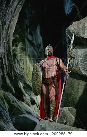 Full length vertical shot of a medieval warrior looking around in a dark forest carrying his shield and a spear wearing a cape and a helmet.