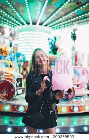 Adorable cute pretty woman stands in middle of amusement park in front of ride with bright colours positive and cheerful eats pink cotton candy floss happy and optimistic