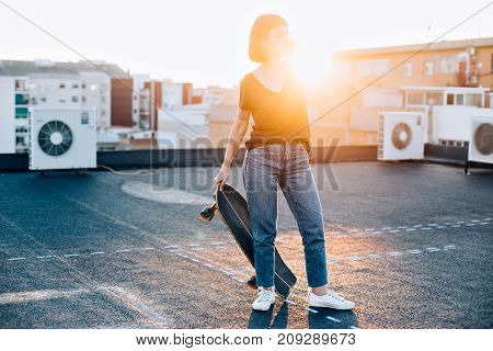 Trendy fashion teenager with lonboard or skateboard stands on top of rooftop during sunset calls friends on smartphone