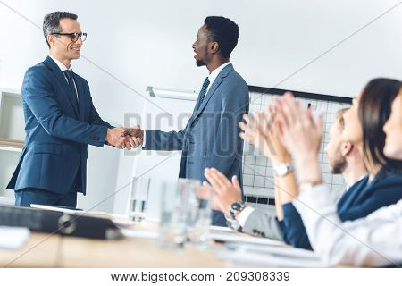 businessman shaking hands in conference hall with partners sitting at table and claping hands