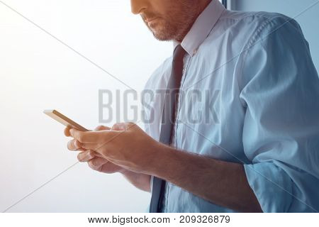 Businessman standing next to office window and texting. Adult caucasian business person in white shirt with tucked rolled up sleeves using mobile phone to send sms message.