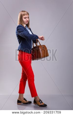 girl in red pants and with leather bag looking at watch over gray background