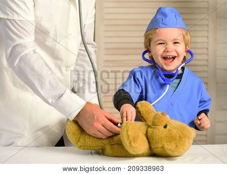 Health and childhood concept. Kid with happy face plays doctor. Male hand holds bear and boy in medical gown holds stethoscope on wooden background. Little assistant examines teddy bear