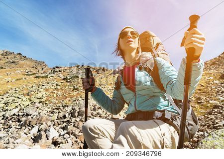 Hiker girl in sunglasses down jacket and hat with a backpack and mountain equipment with tracking sticks in her hands looks at the beautiful view of a high mountain lake in an archipelago in the northern Caucasus. Karachay-Cherkess Republic. The concept o