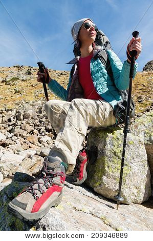 A tourist girl wearing sunglasses down jacket and hat with a backpack and mountain equipment with handles for tracking in her hands is sitting and relaxing looking at the beautiful view of the high mountain lake in the North Caucasus. Karachay-Cherkess Re