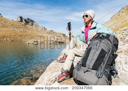 A tourist girl wearing sunglasses down jacket and hat with a backpack and mountain equipment with handles for tracking in her hands is sitting and relaxing looking at the beautiful view of the high mountain lake in the North Caucasus. Karachay-Cherkess Re