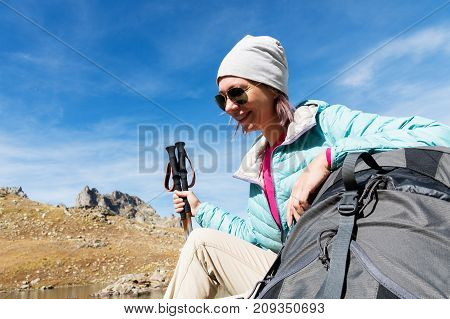 A tourist girl wearing sunglasses down jacket and hat with a backpack and mountain equipment with handles for tracking in her hands is sitting and relaxing looking at the beautiful view of the high mountain lake in the North Caucasus. Karachay-Cherkess Re