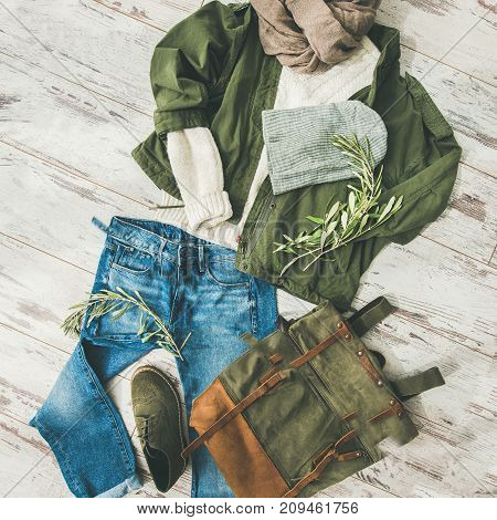 Flat-lay of Fall lady' s clothes outfit. Ecru colored knitted woolen sweater, parka, beige scarf, denim jeans, haki suede boots, backpack, cap over parquet background, top view, square crop