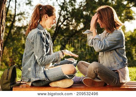 Two pretty girls clap their hands while sitting on wooden bench in park outdoor