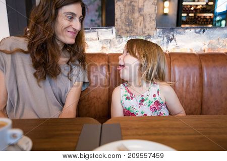 Funny tender scene. Four years age blonde happy girl sticking her tongue out to smiling woman mother sitting in brown leather sofa at restaurant