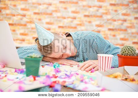 The man in the cap on his head fell asleep in the office at the desk after the party.
