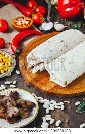 Vegetarian burrito on wooden board over black table surrounded by ingredients