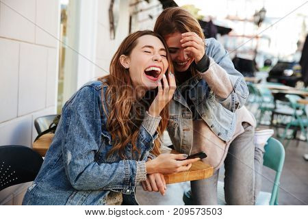Two overjoyed female best friends in jeans wear laughing while sitting at cafeteria