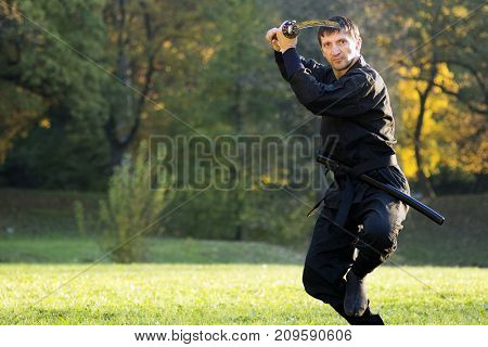 Ninja in black kimono is practicing martial arts with sword outdoors.