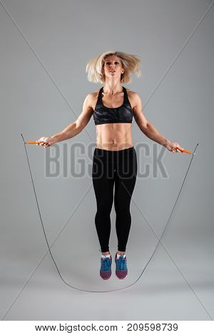 Full length portrait of a concentrated muscular adult sportswoman jumping on a skipping rope isolated over gray background