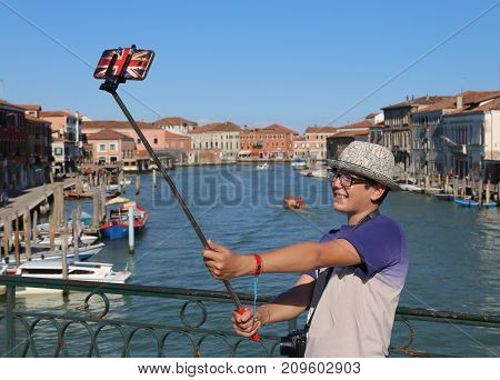 HAppy boy with glasses and hat makes a selfie with the smartphone on the bridge of the island of Murano near Venice in Italy