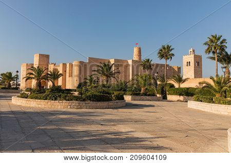 Stone walls of old Ribat fortress and minaret of the Grand Mosque through the lush palms of local garden. Ribat in Monastir is nearly 1200 years old in Monastir. Tunisia.