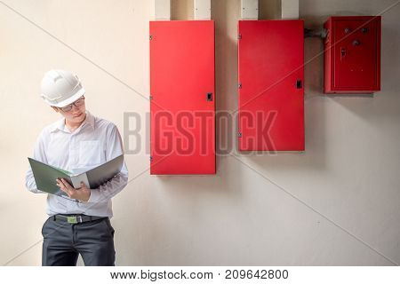 Young Asian Electrical Engineer holding files while wearing a personal protective equipment safety helmet at construction site with red electric control panel. Engineering Maintenance concept.