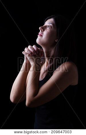Faithful woman praying in worship to god looking up in hope, with hands folded and religious fervor on a black background. Concept for religion, faith, prayer and spirituality.