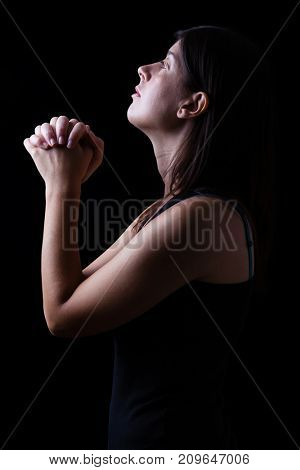 Faithful woman praying in worship to god looking up in hope, with hands folded and religious fervor on a black background. Concept for religion, faith, prayer and spirituality.