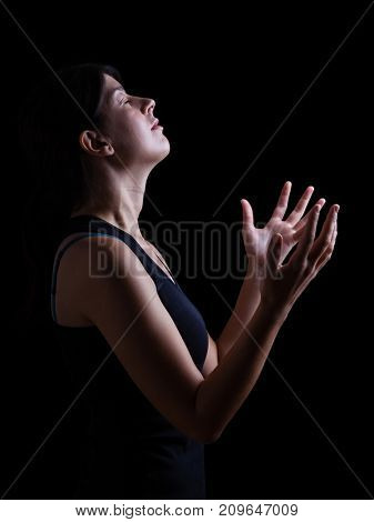 Low key of a faithful woman praying and feeling the presence or being touched by god. Arms outstretched in worship, head up and eyes closed in religious fervor. Black background. 