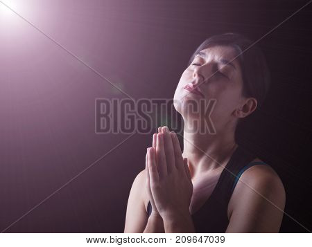 Faithful woman praying under a divine or celestial light and feeling the presence or being touched by god. Hands folded in worship, head up and eyes closed in religious fervor. Black background. 