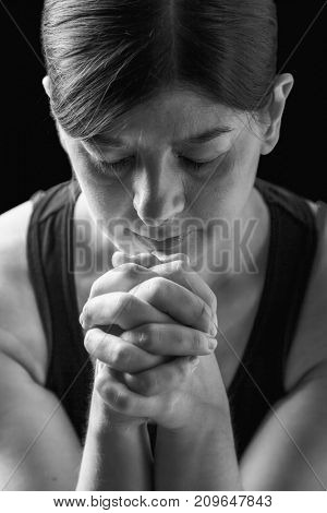 Low key portrait of a faithful woman praying, hands folded in worship to god, head down and eyes closed in religious fervor, on a black background. Concept for religion, faith, prayer and spirituality