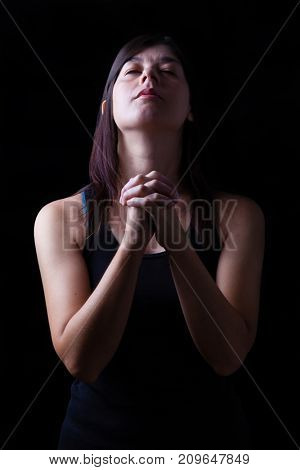 Faithful woman praying, hands folded in worship to god with head up and closed eyes in religious fervor, on a black background. Concept for religion, faith, prayer and spirituality.