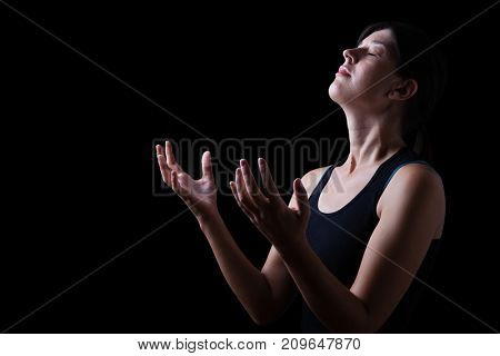 Low key of a faithful woman praying and feeling the presence or being touched by god. Arms outstretched in worship, head up and eyes closed in religious fervor. Black background. 