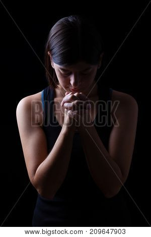Faithful athletic woman praying, with hands folded in worship to god, head down and eyes closed in religious fervor, on low key black background. Concept for religion, faith, prayer and spirituality