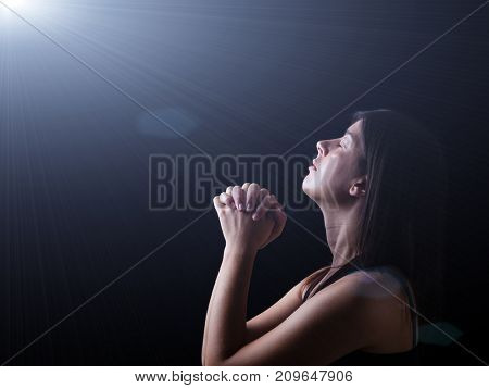Close up of a faithful woman praying under a divine or celestial light, hands folded in worship, head up and closed eyes in religious fervor. Concept for religion, faith, prayer and spirituality.