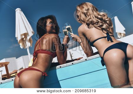 Spending carefree time poolside. Rear view of two stunning young women in bikini enjoying cold drinks while leaning on the poolside outdoors