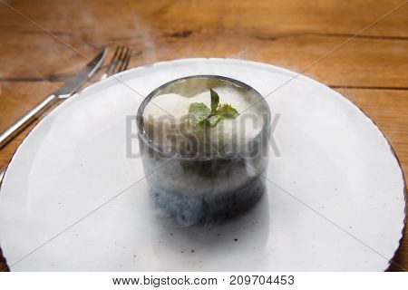 ice cream sweet dessert on a white plate on a wooden table in an Italian retor. Ingredients white ice cream with mint and cocoa in a transparent tequal glass with smoke as per type.