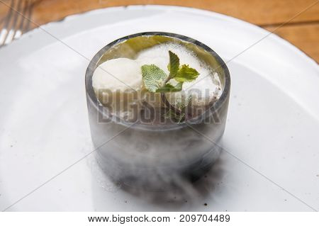ice cream sweet dessert on a white plate on a wooden table in an Italian retor. Ingredients white ice cream with mint and cocoa in a transparent tequal glass with smoke as per type.