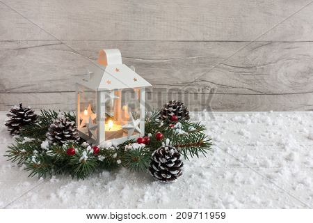 White Christmas lantern with lit candle on fir tree twigs decorated with holly berries pine cones and snowflakes.