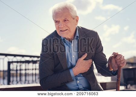 Wincing in pain. White-haired elderly man holding a cane and touching his chest because of suffering from angina