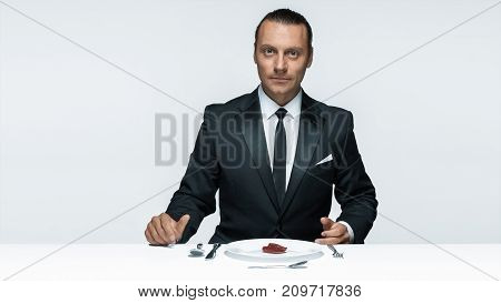 Portrait of a young man in image of maniak with knife, fork and and a piece of raw meat on a plate. Man in suit. The image in the style of Halloween