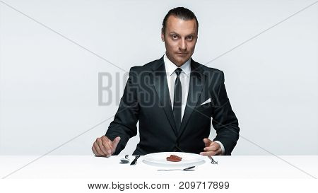 Portrait of a young man in image of maniak with knife, fork and and a piece of raw meat on a plate. Man in suit. The image in the style of Halloween