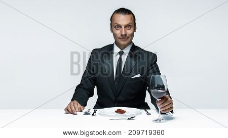 Portrait of a young man in image of maniak with knife, fork and and a piece of raw meat on a plate. Man in suit. The image in the style of Halloween