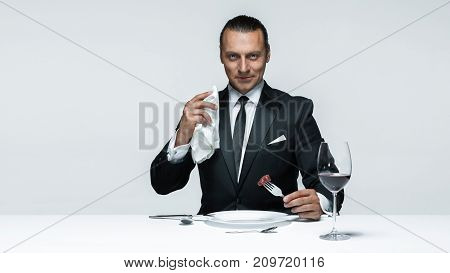 Portrait of a young man in image of maniak with knife, fork and and a piece of raw meat on a plate. Man in suit. The image in the style of Halloween