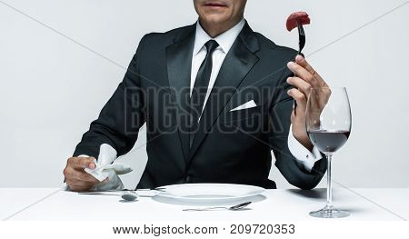 Portrait of a young man in image of maniak with knife, fork and and a piece of raw meat on a plate. Man in suit. The image in the style of Halloween