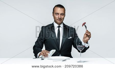 Portrait of a young man in image of maniak with knife, fork and and a piece of raw meat on a plate. Man in suit. The image in the style of Halloween