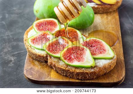 Process of Drizzling Honey with Wood Dipper on Wholegrain Bran Rye Toast with Cream Cheese or Ricotta and Fresh Ripe Figs. Cutting Board. Close Up