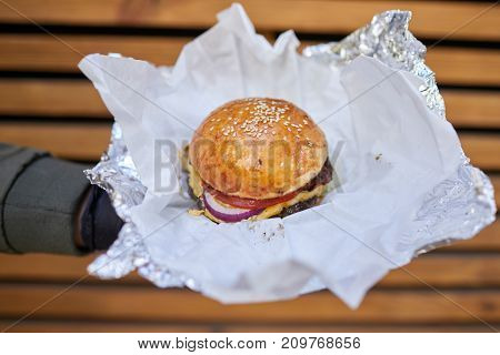Fresh juicy burger with beef, cheese, tomato, salad, majo, onion in male hand in a black gloves on a wooden background. Close-up of burger. Food concept.