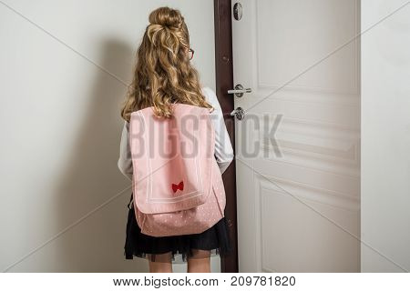 Cute junior schoolgirl with blond hair going to school standing next to door - Back view