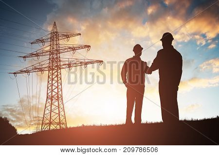 Silhouette Of Two Engineers Shaking Hands With Electricity Pylon Against Dramatic Sky
