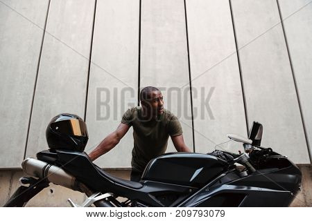 Portrait of a serious young afro american man leaning on a motorcycle and looking away outdoors